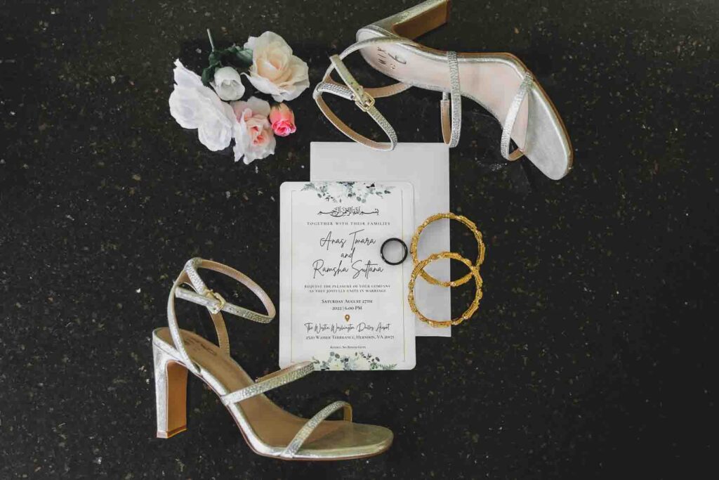 Wedding flat lay with brides shoes, invitation, and wedding rings with flower accents against a black backdrop.