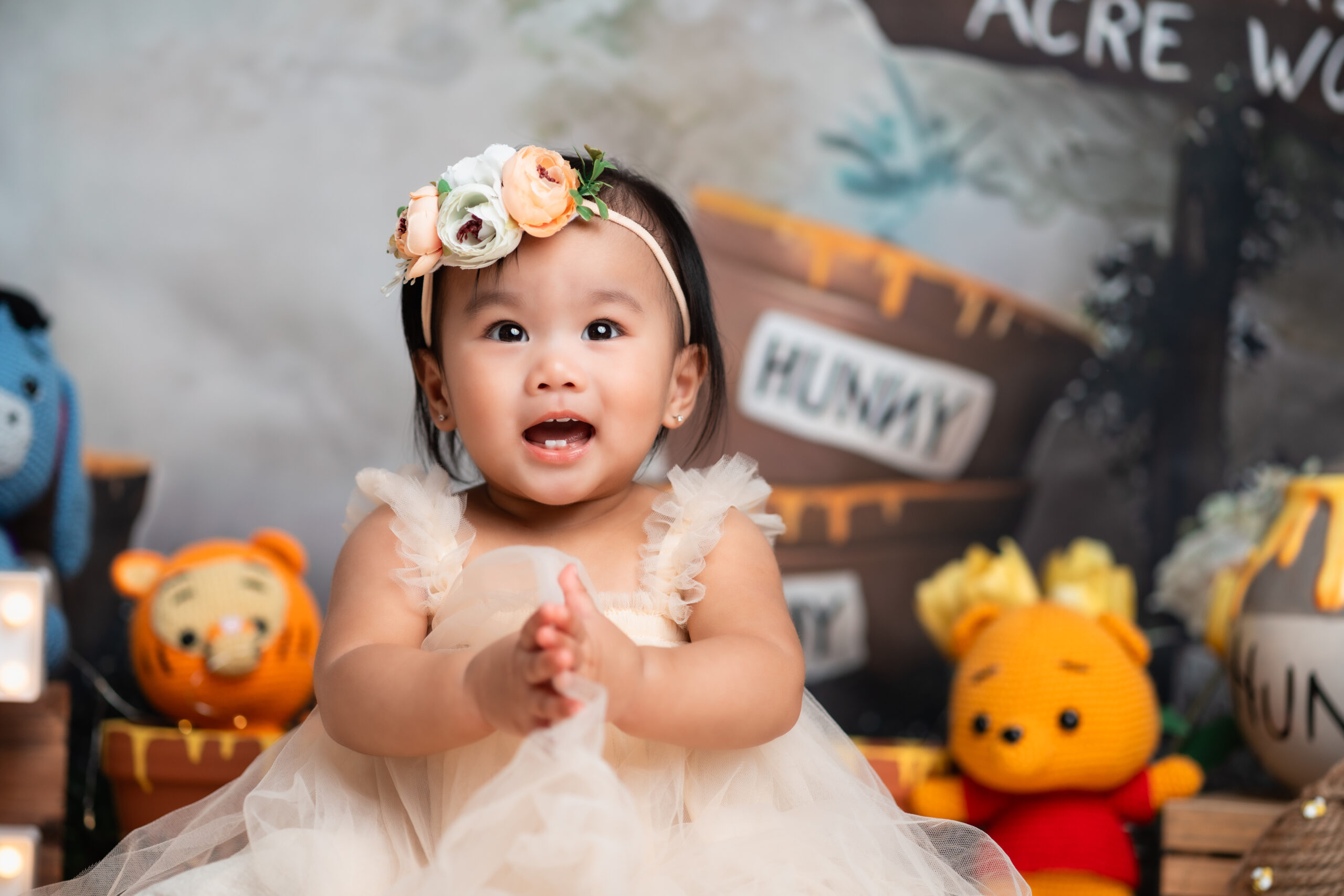 A joyful baby girl clapping hands and sitting in front of a 'Hundred Acre Wood' sign and Winnie the Pooh character stuffed toys. She wears a delicate cream-colored tulle dress and a floral headband. In the foreground, a lit-up marquee sign spells 'ONE' with a red balloon floating above it.