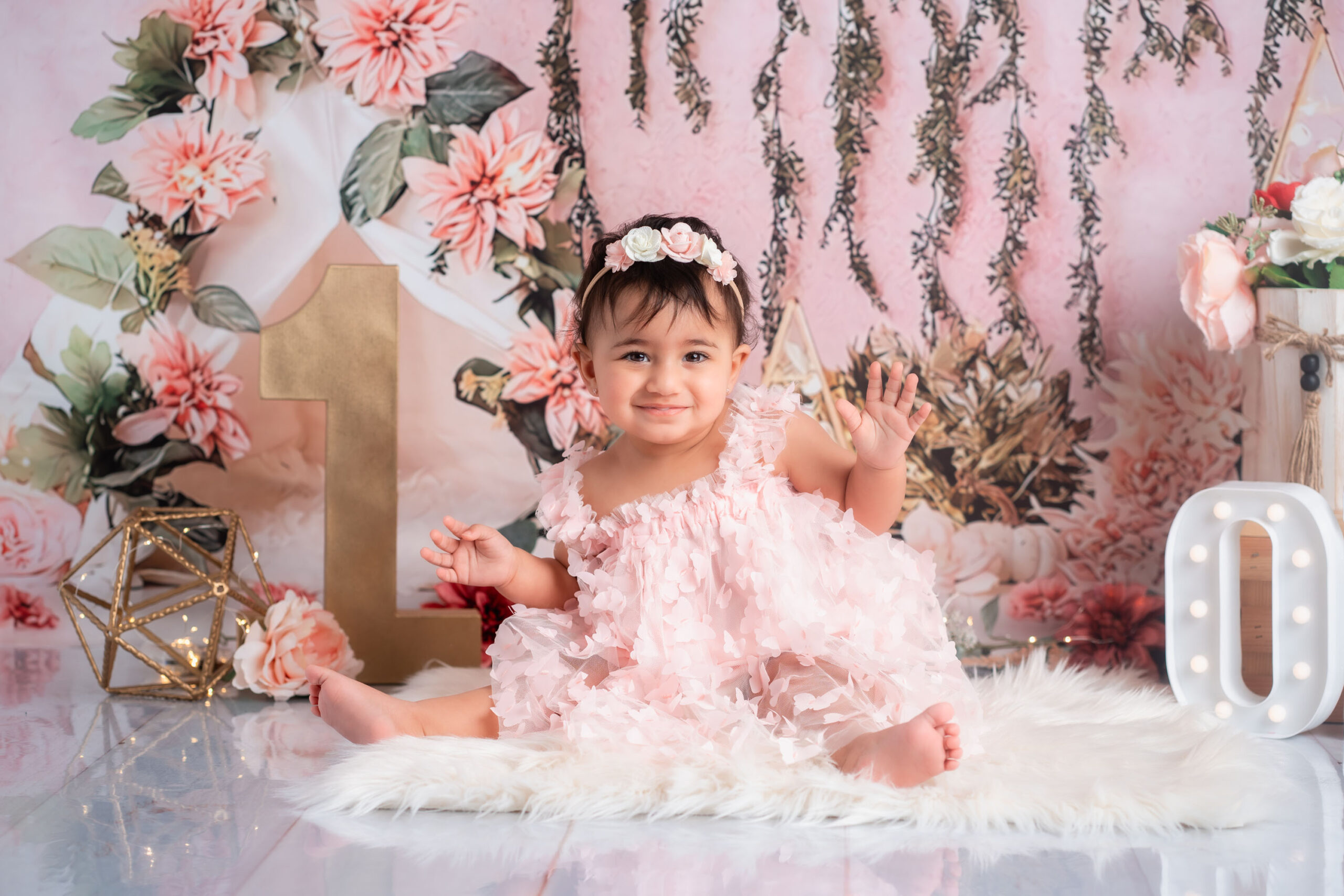 A happy baby girl celebrating her first birthday in front of a pink and white floral background for her cake smash.