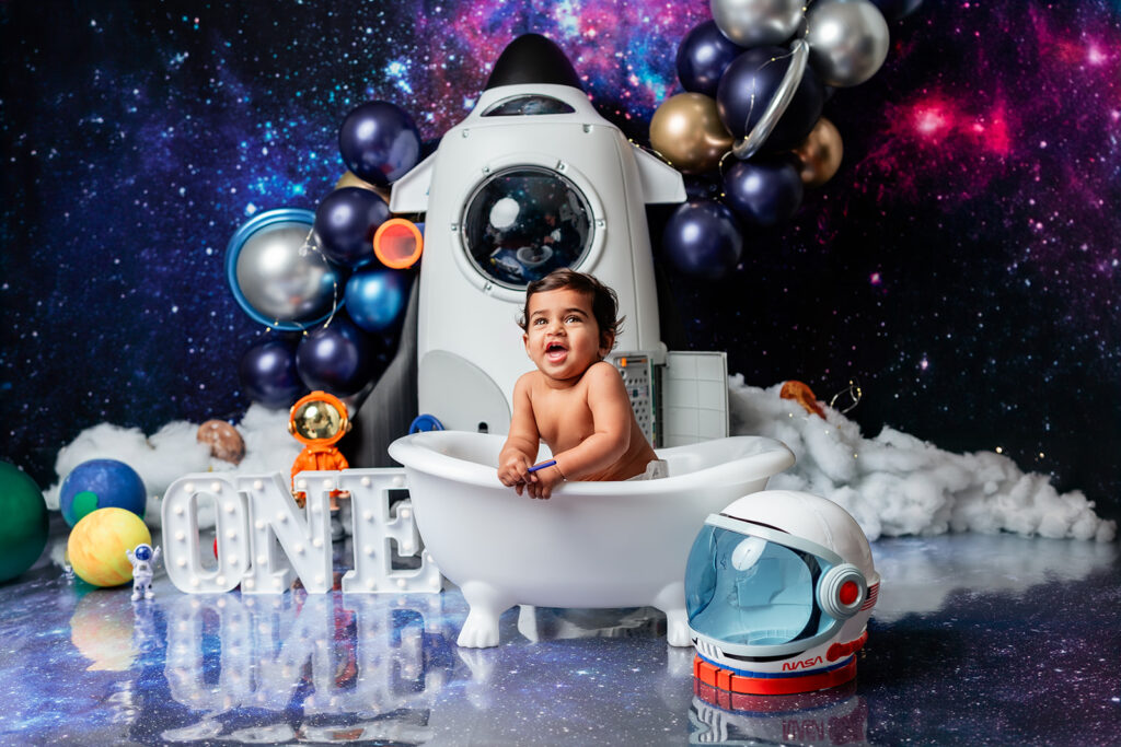 Baby Druv sitting in a mini bathtub with a space-themed backdrop featuring blue and silver balloons and styrofoam planets.