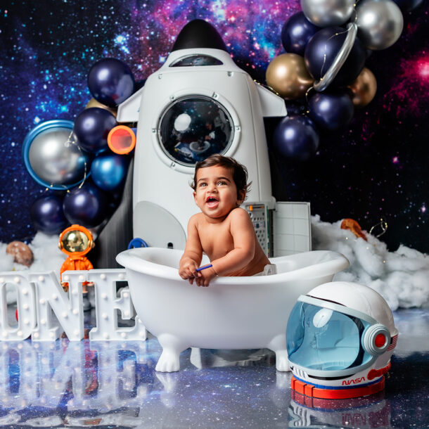 Baby Druv sitting in a mini bathtub with a space-themed backdrop featuring blue and silver balloons and styrofoam planets.