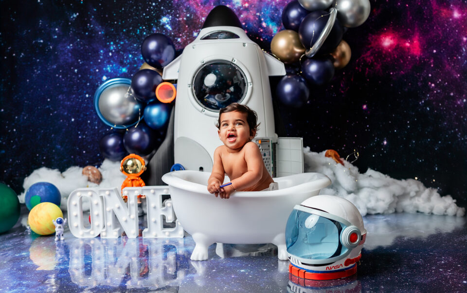 Baby Druv sitting in a mini bathtub with a space-themed backdrop featuring blue and silver balloons and styrofoam planets.